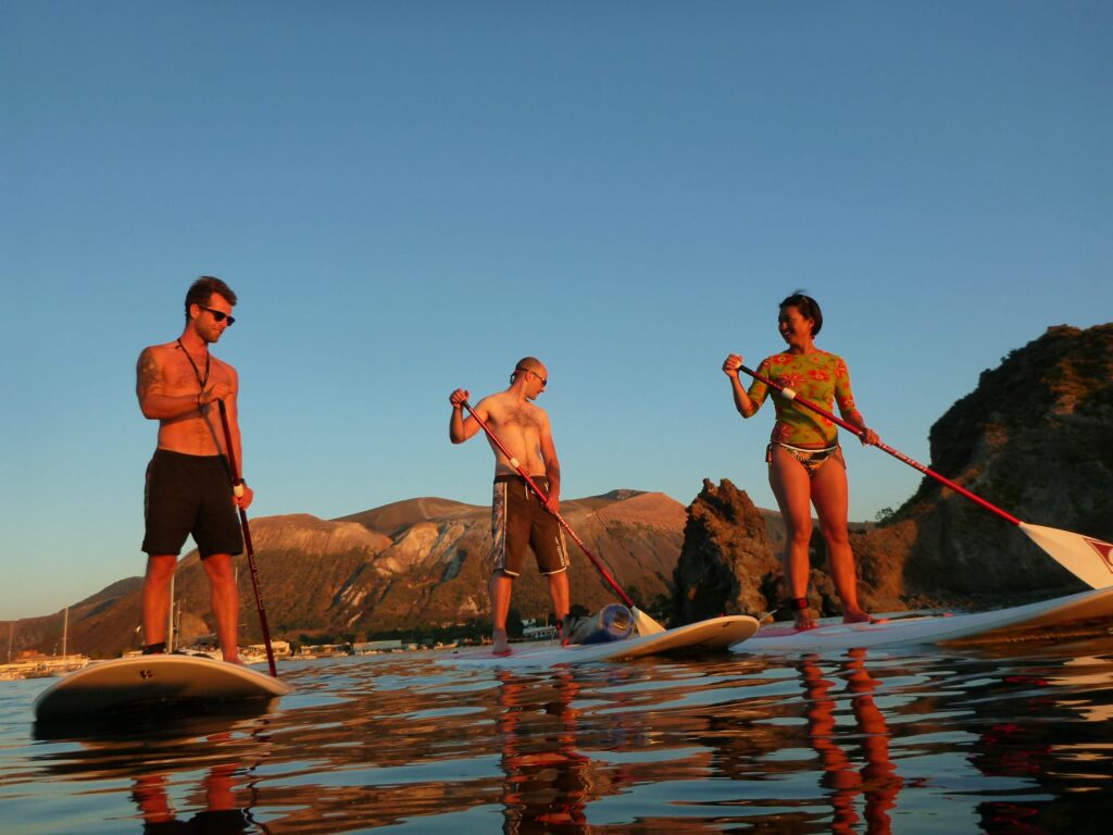 Coppia che partecipa a un'escursione guidata in stand up paddle all'alba intorno a Vulcanello, isola di Vulcano, con il sole che sorge all'orizzonte.