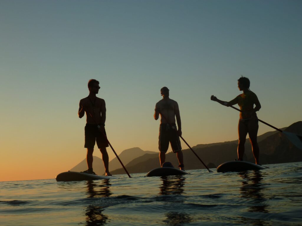 Two people on stand up paddle boards at dawn near Lipari or Vulcano coast with volcanic rocks and calm sea